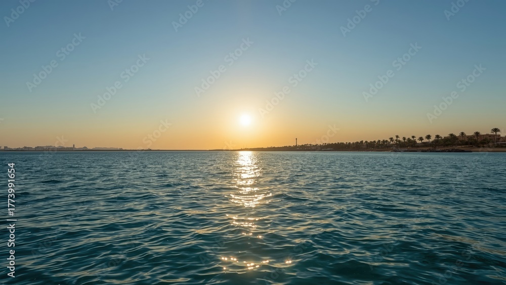 Fototapeta premium A sunset over the water with the horizon, showing a calm sea and a distant shoreline with palm trees. Idyllic scene at dusk or dawn.