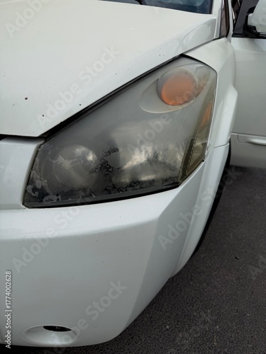 Close-up of a severely oxidized, yellowed, and peeling car headlight on a white vehicle, illustrating the need for restoration, repair, or replacement due to damage and wear.