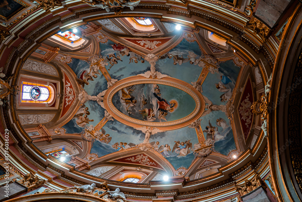 Obraz premium A Baroque dome in Mdina, Malta is seen from below, with pastel blue frescoes of angels and saints, gilded stucco, stained glass windows, and warm light in a wide angle view.