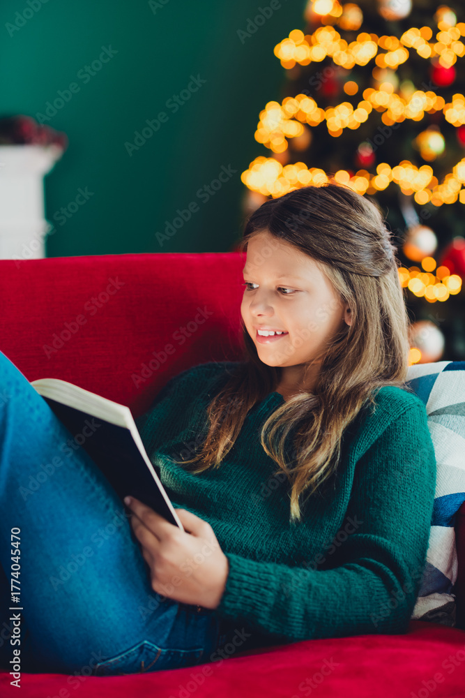 Fototapeta premium A funny cozy scene of a girl reading a book beside a glowing Christmas tree during winter holidays at home
