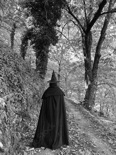 Black and white photo of a woman dresses as a witch in a cloak walking in the forest