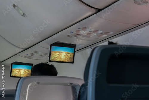Interior second-class aircraft cabin with seated unrecognizable passengers and shared overhead screens displaying flight information during the journey over the yellowish sands of the Sahara desert.