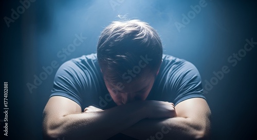 A person with arms crossed, head bowed, in a dark setting with moody lighting