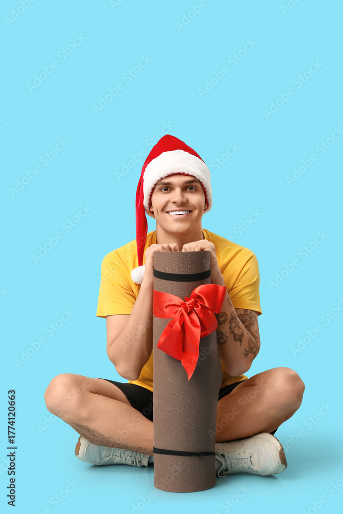 Obraz premium Young happy man in Santa hat sitting with aerobic mat on blue background