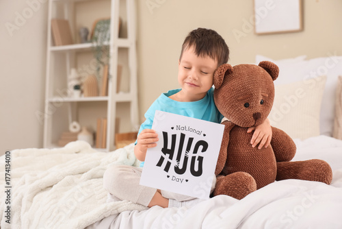 Cute little boy holding paper with text NATIONAL HUG DAY and toy bear in bedroom