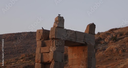 Warm evening light bathes a weathered stone gateway at Persepolis as two pigeons perch on the blocks. A carved Faravahar recalls the Achaemenid empire—quiet endurance in Iran’s desert foothills.