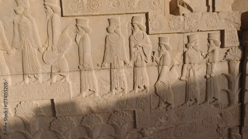 Tracking along an Achaemenid stone relief at Persepolis, Iran: bearded courtiers in procession beneath carved rosettes, crisp sunlight and ancient textures evoking empire, ritual, timeless heritage.