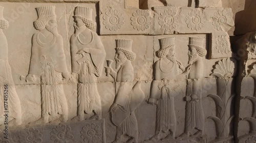 Tracking along an Achaemenid stone relief at Persepolis, Iran: bearded courtiers in procession beneath carved rosettes, crisp sunlight and ancient textures evoking empire, ritual, timeless heritage.