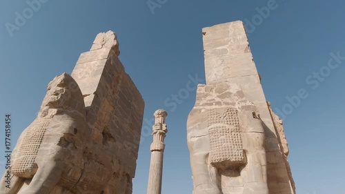 Wallpaper Mural Low-angle tracking shot between the colossal guardian bulls of the Gate of All Nations at Persepolis, Iran, revealing fluted columns and Achaemenid stonework under a clear blue sky. Torontodigital.ca