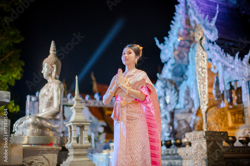 woman wearing a traditional thai dress smiling and greeting with wai gesture in front of wat sri suphan temple during yi peng and loy krathong festival night