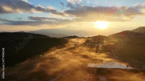 Aerial clip of mountain valley mist in the morning with the rising sun breaking through the clouds to cast its light onto the mist, turning it a golden colour with long shadows.