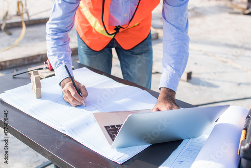 Construction worker wearing a working suit with helmet, holding a pen and looking at the papers with architectural site blueprint placed on the table.
