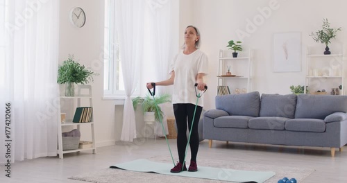Healthy physically fit adult senior old woman standing on a sports mat in the living room at home and doing arm strength exercises with an elastic fitness band with handles