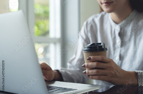 Business woman hand holding a paper cup of coffee during working on laptop computer at home office or coffee shop. taking a break, freelance at work, people lifestyle