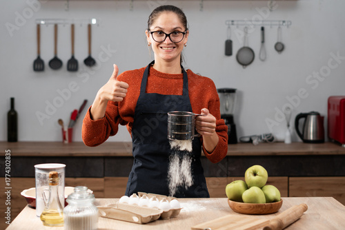 Woman housewife in apron and eyeglasses sifting flour making dough for cake showing like gesture thumb-up while cooking food in the kitchen at home.