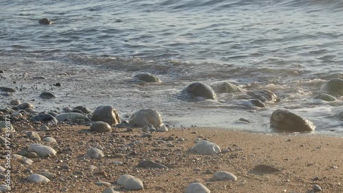 Powerful ocean waves crash and foam against the rough, sun-drenched rocky and pebble-covered shoreline.