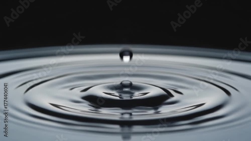 A close-up macro shot captures a single water droplet falling into a perfectly still pool, creating concentric ripples that spread outwards across the dark, reflective surface
