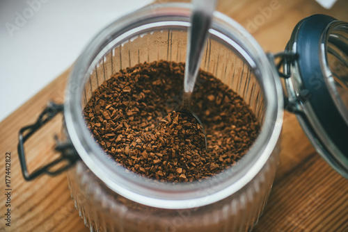 A spoon dipped into a glass jar filled with instant coffee granules on a wooden table, captured in warm natural light.