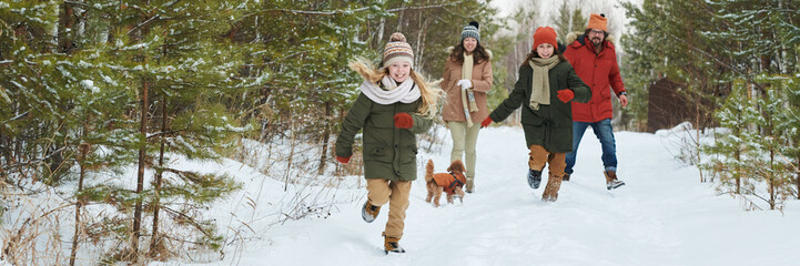 Two Caucasian kids running through snowy forest path with small brown dog while Caucasian woman and Caucasian man following behind, all wearing winter clothing, enjoying outdoor activity