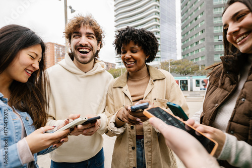 Happy young group of diverse friends having fun using mobile phones at city street, texting messages or watching social media content on cell app