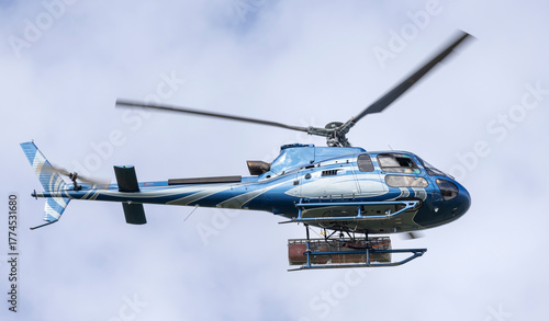A blue helicopter flies with cargo under the clouds
