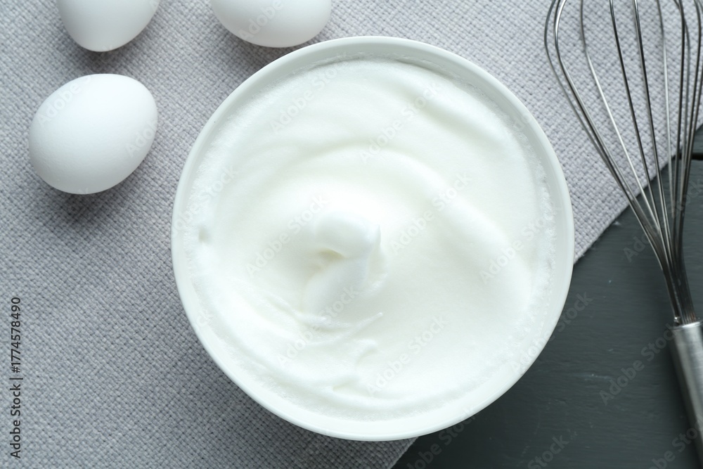Fototapeta premium Whipped whites in bowl, whisk and eggs on grey wooden table, flat lay