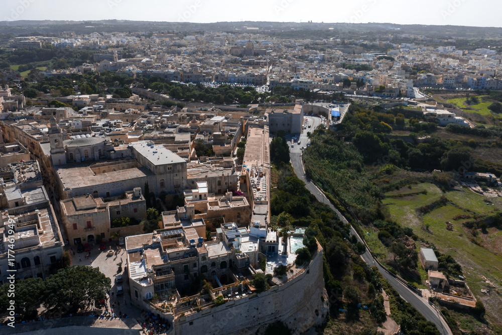 Fototapeta premium Aerial view shows Mdina, Malta, with honey colored walls, narrow streets, bastions, and main gate, midday light, visitors by the walls, Rabat and fields beyond