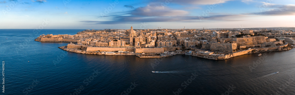 Obraz premium Aerial view of Valletta, Malta at sunset shows limestone bastions, Fort St. Elmo, the basilica dome, and cathedral spire as boats cross Marsamxett Harbour.