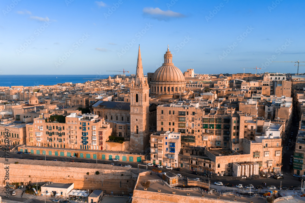 Fototapeta premium Aerial view of Valletta, Malta at sunset, basilica dome and cathedral spire rise above limestone streets and bastions, golden hour light warms sandstone beside the sea.