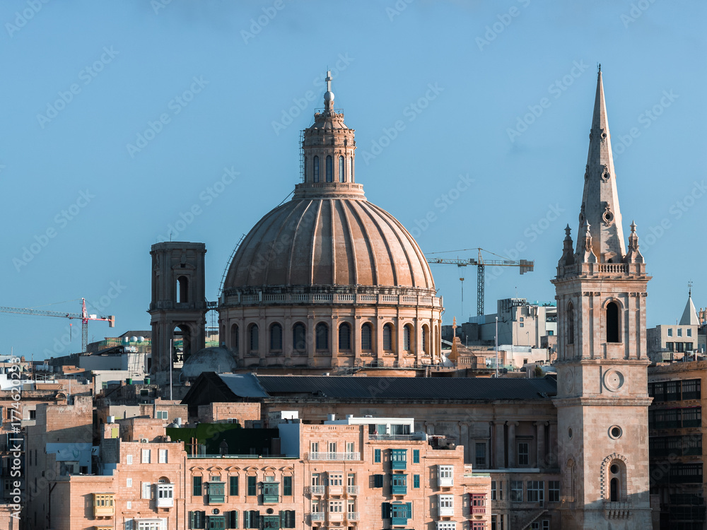 Fototapeta premium Valletta, Malta cityscape shows basilica dome and pro cathedral spire above limestone buildings, rooftop terraces, and cranes in clear daylight from an elevated vantage.