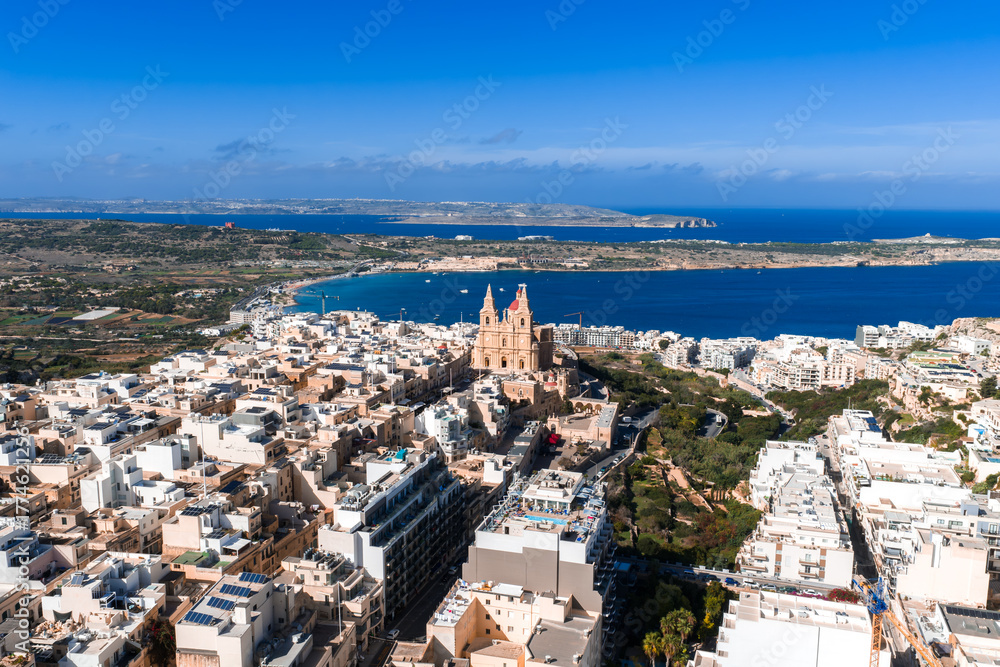 Fototapeta premium Aerial view shows Mellieha, Malta, with the red sandstone parish church above white buildings, Mellieha Bay, and Comino and Gozo on the horizon in bright midday light.