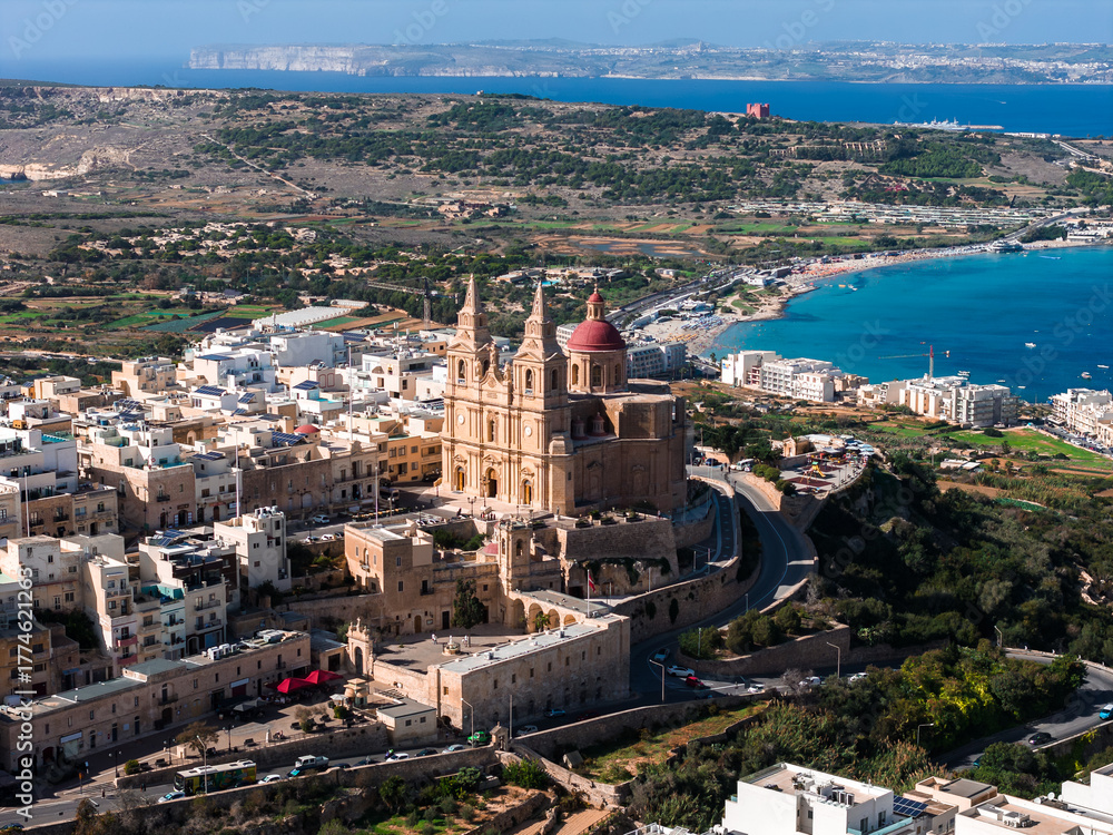 Fototapeta premium Aerial view of the Parish Church of Mellieha with twin spires and red dome above limestone buildings, Mellieha, Malta. Mellieha Bay, Comino, and Gozo appear in clear daylight.