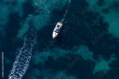 Foto Aerial view shows a white motor yacht cutting across deep blue water off Sliema, Malta, foamy wake curving left, turquoise shallows and rock patches visible at midday