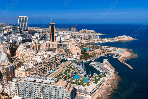 Aerial view of Sliema and St. Julian's, Malta, shows Portomaso Marina, the cylindrical Portomaso Business Tower, rocky peninsula, bays, pools, promenades, and beaches at midday.