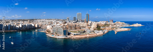 Fototapeta Aerial panorama of Sliema, Malta shows rocky shore, promenade, clear bays with boats, limestone blocks, glass towers, cranes, seawalls, lidos in bright midday light