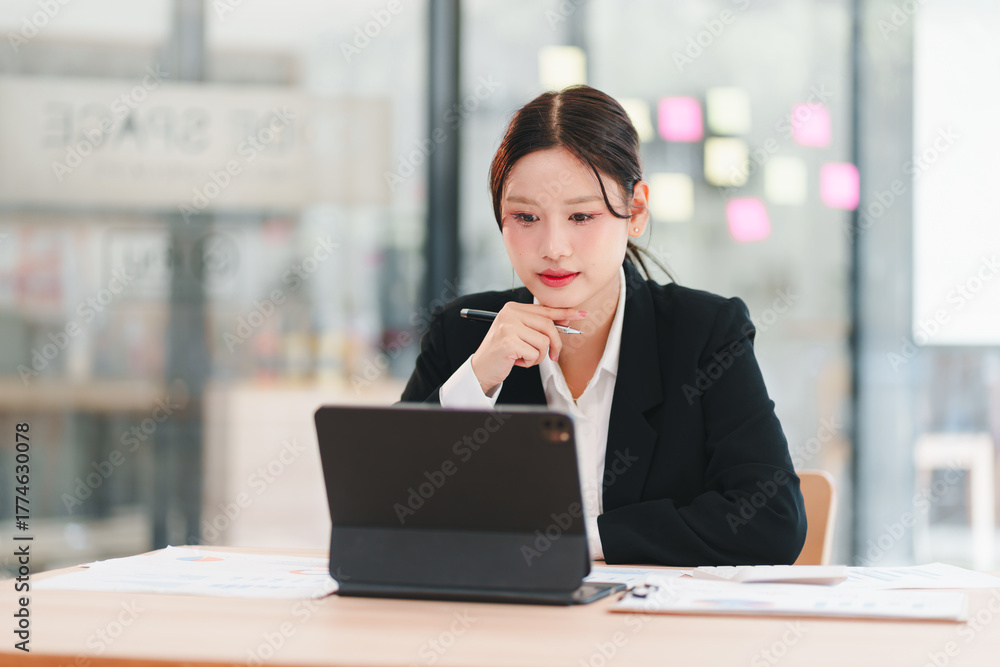 Fototapeta premium Young woman in business suit working on tablet and reviewing documents with focused expression