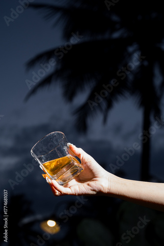 A hand holding a glass of golden drink against dark sky and palm tree silhouette, representing relaxation and evening tropical atmosphere.
