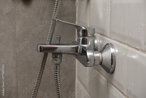 Close-up of a shiny chrome shower faucet with attached hose, mounted on a white tiled wall in a modern bathroom setting.

