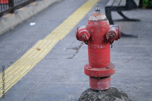 Weathered red fire hydrant installed on a city sidewalk next to yellow tactile paving for visually impaired pedestrians, with benches nearby.
