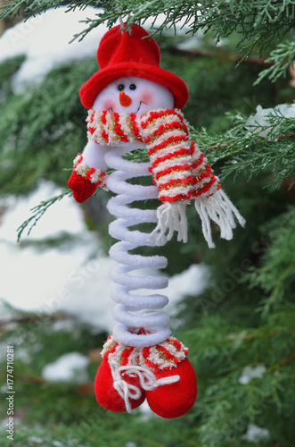 Cute snowman toy in red hat , scarf and shoes hanging on a snowy branch of a snowy fir tree. Closeup outdoors photo. Christmas greeting concept. Free copy space. 
