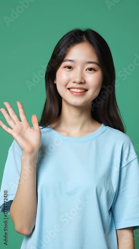 Beautiful Asian girl in a light blue casual T-shirt smiling warmly and waving at the camera, standing against a solid green background, soft daylight, realistic style, friendly atmosphere.
