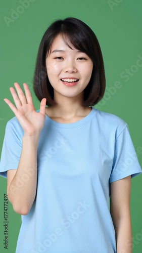 Beautiful Asian girl in a light blue casual T-shirt smiling warmly and waving at the camera, standing against a solid green background, soft daylight, realistic style, friendly atmosphere.
