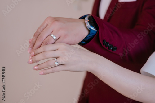 The bride's left hand holding the wedding ring is holding the groom's left hand who is also wearing a wedding ring.