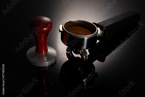 Espresso machine portafilter handle with compacted coffee and wooden tamper over gradient black background with reflection.