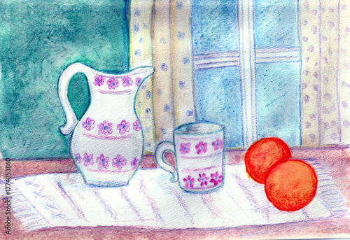 Two oranges, a cup and a jug with a floral pattern standing on a table by a window.
