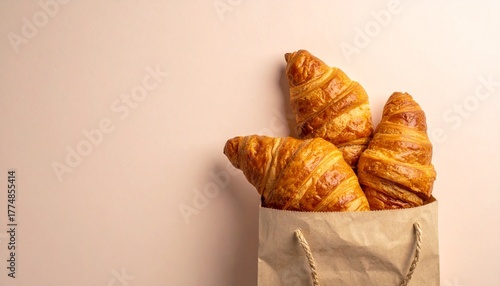 Three golden brown croissants peek out from a crumpled white paper bag, set against a soft peach background