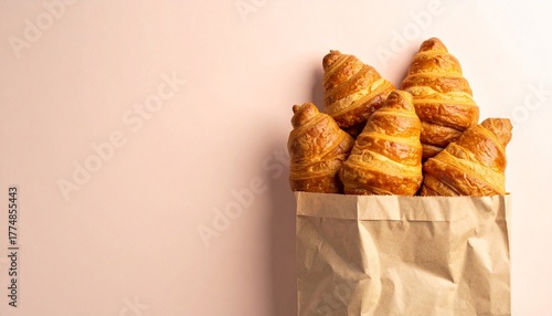 Three golden brown croissants peek out from a crumpled white paper bag, set against a soft peach background