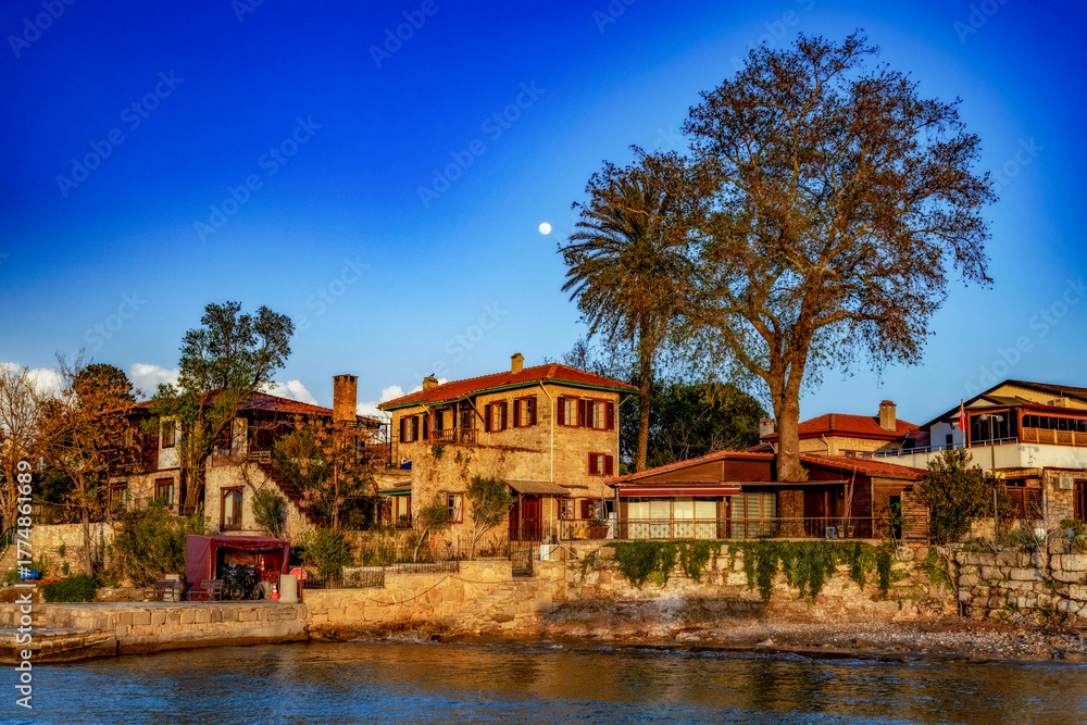 Obraz premium Sunset view from the sea of Side's old town with traditional stone houses, wooden shutters, and a full moon rising over tile roofs. Antalya, Turkey.