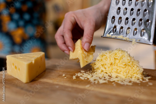 Detailed shot of cheese being grated, Metal grater creating fine cheese curls on wooden surface, Closeup of fingers creating textured cheese shreds for culinary