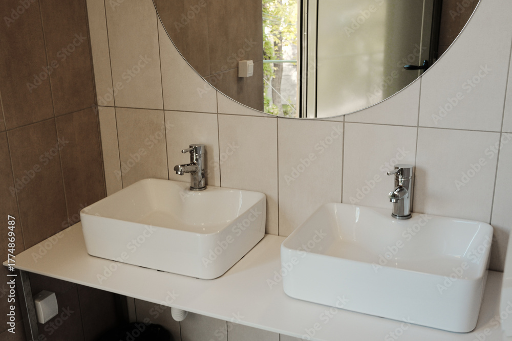 Fototapeta premium Two modern white sinks with chrome faucets and large round mirror inside tiled restroom